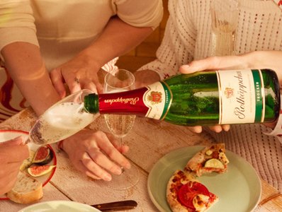 Zwei Hände servieren prickelnden Sekt, während ein kleiner Teller mit Pizza und frischen Feigen im Hintergrund steht.