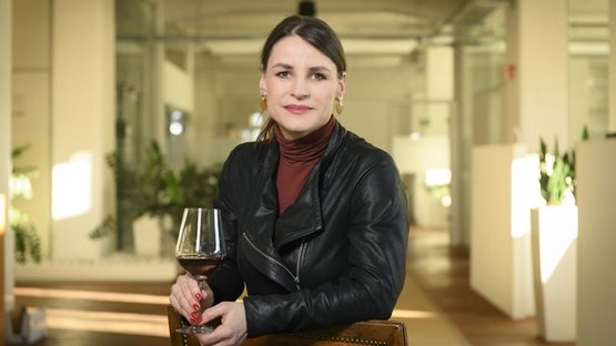 Eine Frau mit einem Glas Rotwein sitzt in einem modernen Raum, umgeben von Pflanzen und hellem Licht.