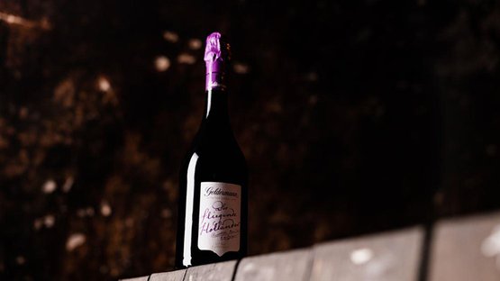 Eine elegante Flasche Wein mit lila Kapsel steht auf einem rustikalen Holzregal vor einem unscharfen, dunklen Hintergrund.