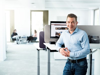 A smiling man in a bright office stands at a standing desk while two women in the background are engrossed in conversation.