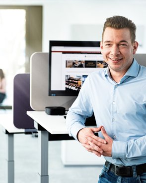 A smiling man in a bright office stands at a standing desk while two women in the background are engrossed in conversation.
