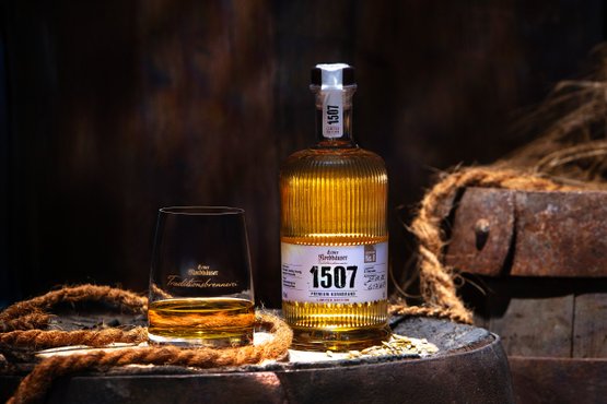 Eine Flache Echter Nordhäuser Traditionsbrennerei 1507 No. 8 und ein Glas stehen auf einem Holzfass vor dunklem, rustikalem Hintergrund.