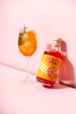Polly-Flasche lehnt an der Wand, daneben ein Cocktailglas mit einem orangefarbenen Aperitf