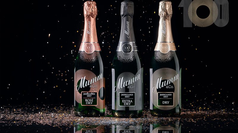 Drei Flaschen Sekt von Mumm & Co. stehen auf einem glänzenden Tisch, umgeben von glitzerndem Konfetti vor schwarzem Hintergrund.