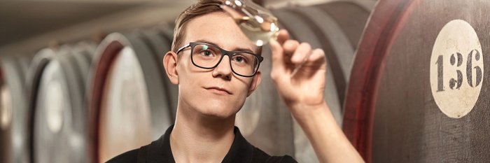 Ein junger Mann mit Brille betrachtet aufmerksam ein Glas Wein in einem Weinkeller, umgeben von Holzfässern.