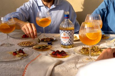 A hand reaches for an olive, while two glasses of orange drinks and a bottle of Canonita stand in the background.