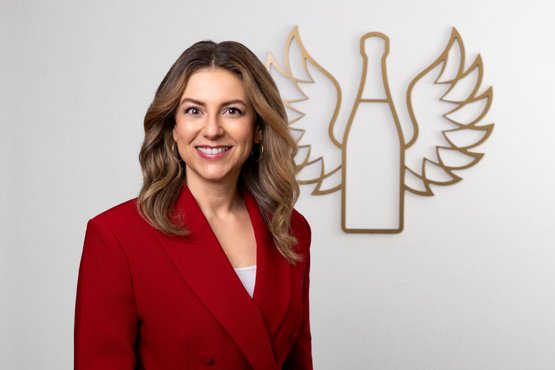 Silvia Wiesner: A woman with long, wavy hair smiles friendly in a red blazer in front of a wall with a bottle logo.