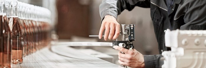 Ein Arbeiter in einem grauen Overall bedient eine Maschine zur Abfüllung von Flaschen mit roséfarbenem Getränk.