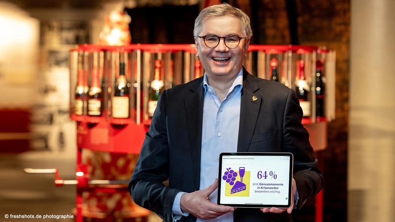 Ein lächelnder Mann hält ein Tablet mit einer Grafik, die besagt, dass 64 % Genussmomente in Krisenzeiten wichtig sind.