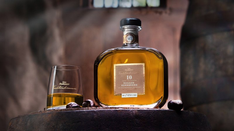 Eine edle Flasche Roggenkornbrand steht auf einem Holzfass, daneben ein Glas mit goldenem Inhalt, umgeben von Kastanien.