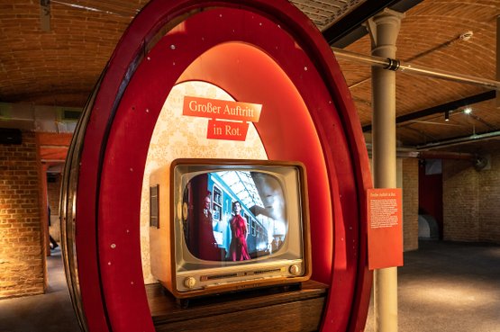 Ein nostalgischer Fernseher in einem roten Rahmen zeigt eine Szene mit einer Frau in einem roten Kleid vor einem Zug.