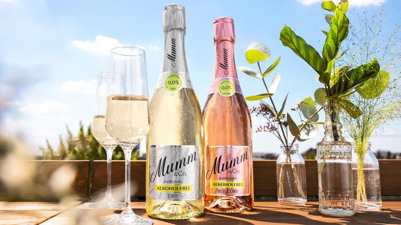 Zwei Flaschen alkoholfreier Sekt stehen auf einem Holztisch, umgeben von frischen Blumen und Gläsern im Sonnenlicht.