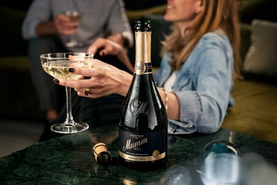 A woman holds a glass of sparkling wine in her hand while a bottle of Mumm Riesling Brut sits on the table.