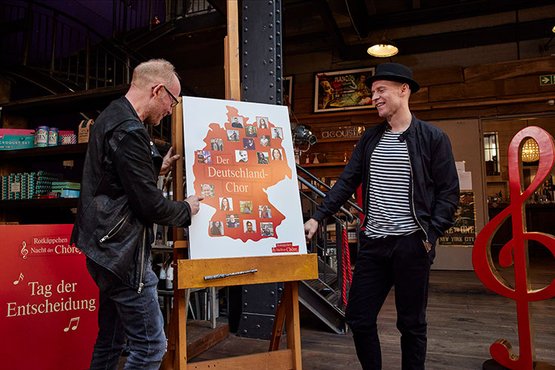 Zwei Männer stehen vor einem Plakat, das den Deutschland-Chor zeigt, während sie miteinander lachen und diskutieren.