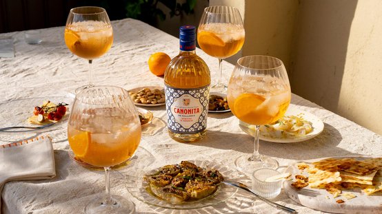 Ein Tisch gedeckt mit Gläsern voller orangefarbener Getränke, umgeben von verschiedenen Snacks und einer Flasche Canonita.