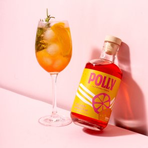 Polly-Flasche lehnt an der Wand, daneben ein Cocktailglas mit einem orangefarbenen Aperitf