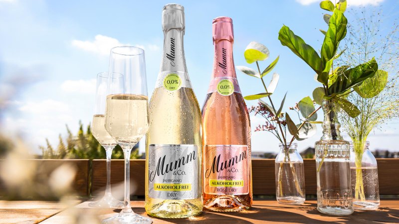 Zwei Flaschen alkoholfreier Sekt stehen auf einem Holztisch, umgeben von frischen Blumen und Gläsern im Sonnenlicht.