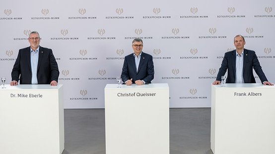 Drei Männer stehen an weißen Tischen, lächeln und präsentieren sich vor einer neutralen Wand mit dem Logo von Rotkäppchen-Mumm.