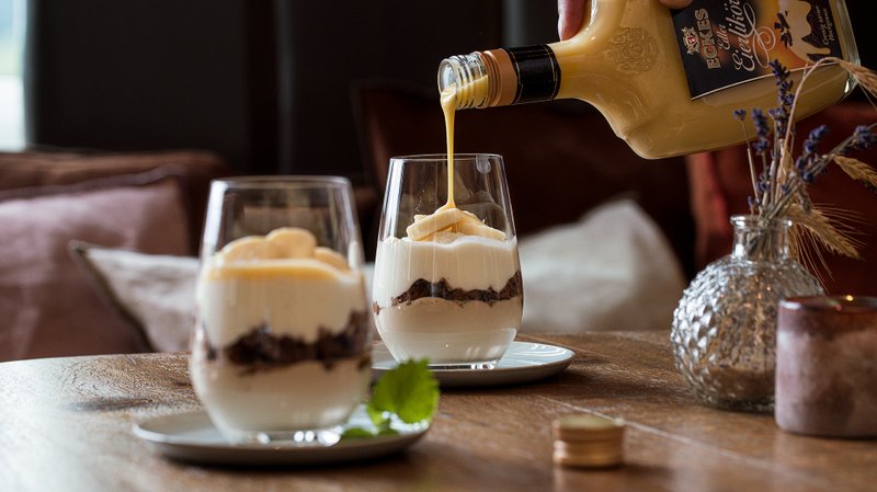 Ein Glas mit Schichtdessert wird mit Eierlikör übergossen, während ein zweites Glas im Hintergrund sichtbar ist.