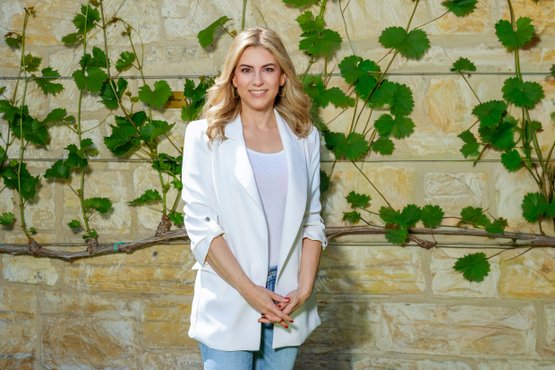 Porträtfoto Silvia Wiesner: Sie trägt einen weißen Blazer und Jeans, während sie entspannt mit gefalteten Händen vor einer natürlichen Kulisse posiert.