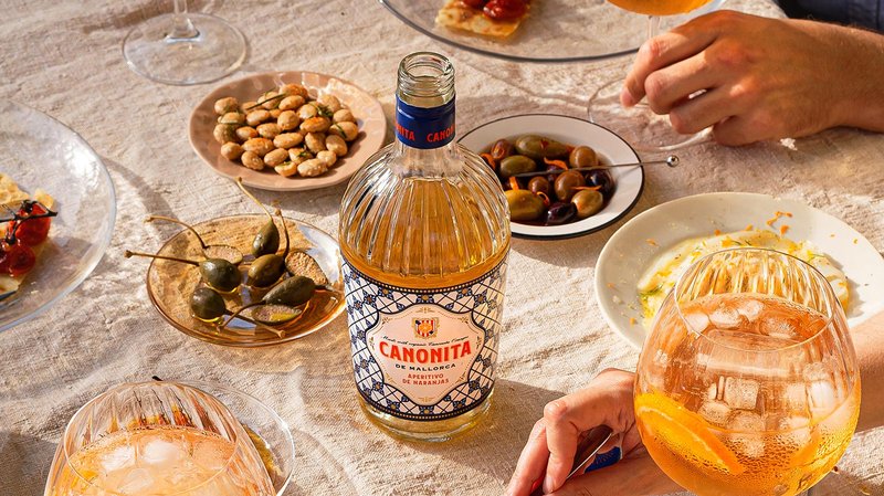 Canonita: Auf einem hellen Tisch liegen verschiedene Leckereien, während eine Hand ein Glas mit einem spritzigen Getränk hält.