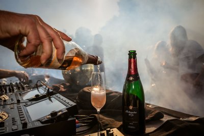 Ein DJ-Pult im Vordergrund, während ein Glas mit sprudelndem Sekt gefüllt wird, umgeben von verschwommenen Feiernden.