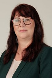 Eine lächelnde Frau mit kurzen, roten Haaren und Brille trägt einen grauen Blazer und steht vor einem hellen Hintergrund.