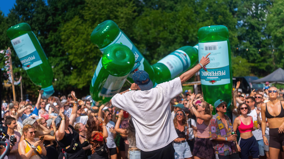 Ein begeisterter Sänger steht vor einer jubelnden Menschenmenge, während riesige grüne Pfeffi-Flaschen durch die Menge getragen werden.