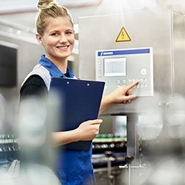 Eine fröhliche Mitarbeiterin in einem blauen Arbeitsanzug bedient eine Maschine und hält einen Clip-Ordner in der anderen Hand.