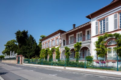 Eltville: An elegant building with a balcony, surrounded by lush greenery and colorful flowers, under a bright blue sky.