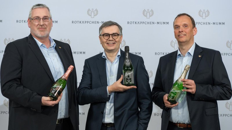 Drei Männer in Anzügen halten Flaschen Sekt, lächeln und stehen vor einem Hintergrund mit dem Logo von Rotkäppchen-Mumm.