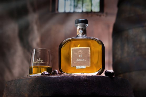 Eine edle Flasche Roggenkornbrand steht auf einem Holzfass, daneben ein Glas mit goldenem Inhalt, umgeben von Kastanien.