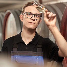 Ein Mann mit Brille und Arbeitskleidung inspiziert ein Glas Wein, um dessen Klarheit und Farbe zu beurteilen.