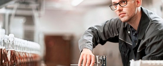 Ein Mann in einem grauen Arbeitsanzug überprüft sorgfältig eine Maschine in einer Produktionsumgebung mit Flaschen im Hintergrund.