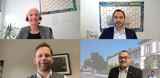 Eine Gruppe von vier Personen in einem virtuellen Meeting, lächelnd und in professioneller Kleidung, mit einem Gebäude im Hintergrund.