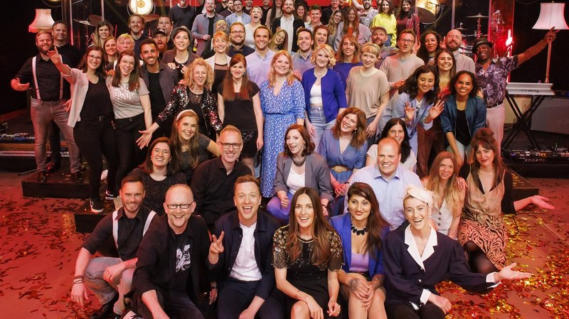 Eine große Gruppe von Menschen posiert fröhlich auf einer Bühne, umgeben von buntem Konfetti und festlicher Beleuchtung.