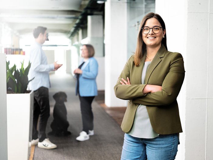 A confident woman in an olive green blazer smiles while standing in a modern office with plants.