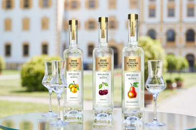 Drei Flaschen Schloss Salem Obstbrand stehen auf einem Tisch, umgeben von eleganten Gläsern und einer grünen Gartenlandschaft.