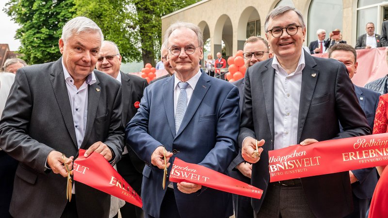 Drei Männer in Anzügen halten eine große Schere und schneiden ein rotes Band durch, während sie lächeln.