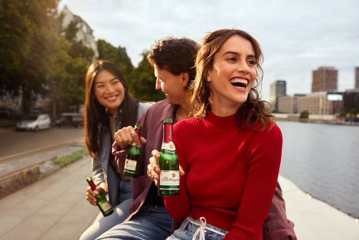 Drei Personen, darunter eine Frau im roten Pulli, sitzen am Wasser und halten Piccolo-Flaschen Rotkäppchen in der Hand.