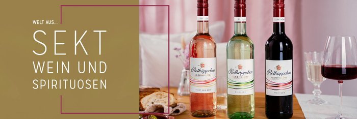 Drei Flaschen alkoholfreier Rotkäppchen-Weine stehen auf einem Holztisch, umgeben von einem Glas Rotwein und einem Dessert.