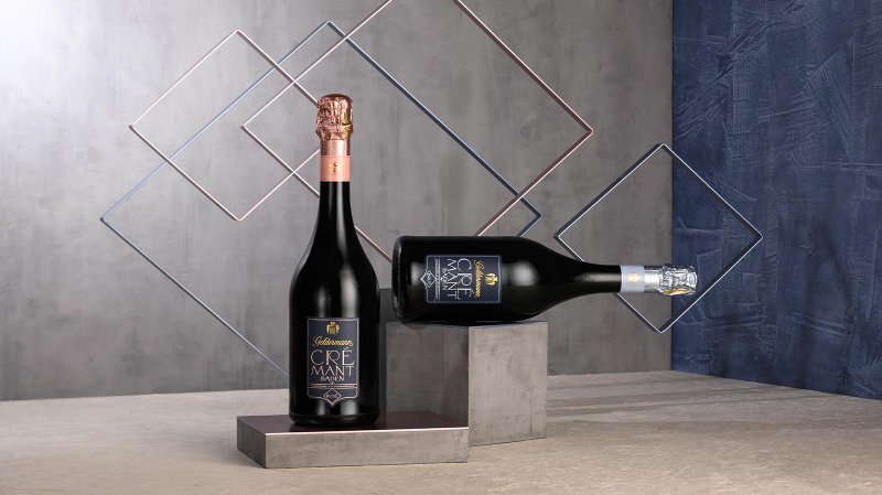 Zwei Flaschen Crémant stehen auf geometrischen Podesten, umgeben von modernen, farbigen Rahmen auf einem neutralen Hintergrund.