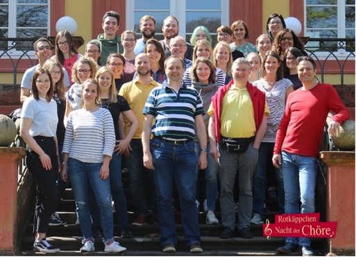 Eine Gruppe von 30 Personen steht fröhlich auf einer Treppe vor einem farbenfrohen Gebäude, umgeben von Bäumen und Tageslicht.