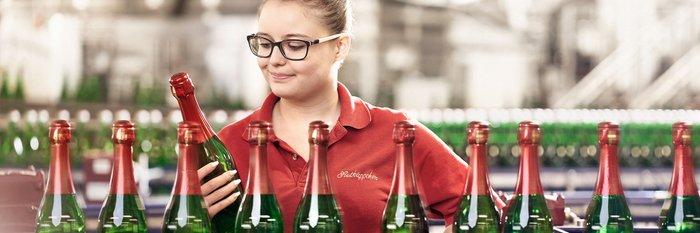 Eine Frau mit Brille betrachtet eine Flasche Sekt in einer Produktionsstätte, umgeben von vielen weiteren Flaschen.