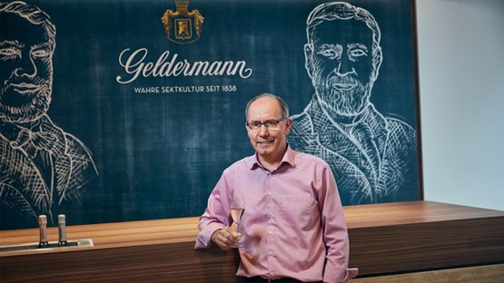 Der Mann mit Brille und rosa Hemd präsentiert stolz ein Glas Sekt vor einer Wand mit dem Schriftzug 'Geldermann'.