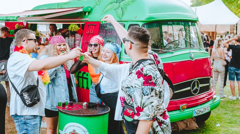 Eine Gruppe junger Menschen hebt ihre Gläser vor einem bunten Foodtruck und feiert fröhlich zusammen.