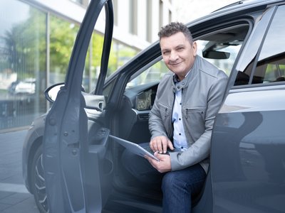 Ein Mann in grauer Jacke sitzt in einer geöffneten Autotür und blickt mit einem leichten Lächeln in die Kamera. In den Händen hält er ein Tablet. Im Hintergrund ist ein Gebäude mit großen Fenstern zu sehen.