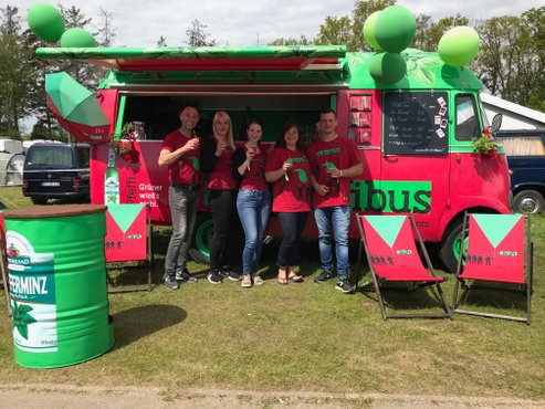 Fünf Personen in roten T-Shirts stehen vor einem bunten Foodtruck, während sie Getränke in der Hand halten und lächeln.