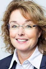 A smiling woman with curly brown hair wears glasses and a dark blazer against a light background.