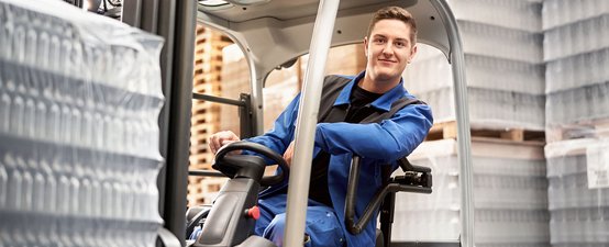 Ein junger Mann in blauer Arbeitskleidung sitzt lächelnd in einem Gabelstapler, umgeben von Paletten und Lagerregalen.